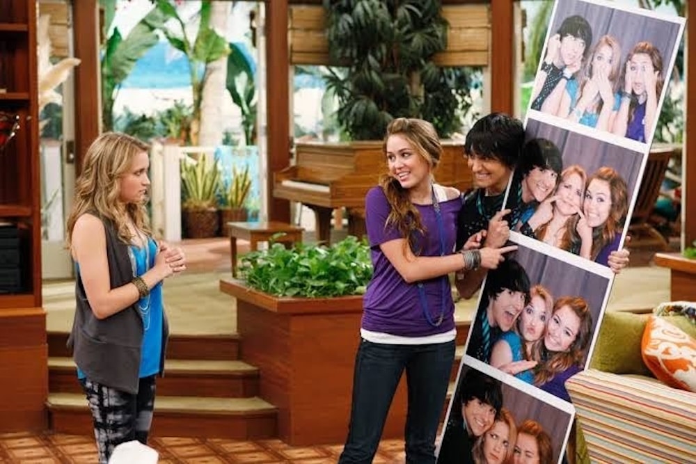 Three teenagers stand in a colorful living room; two excitedly hold a large board with fun photo booth pictures, while the third looks on in surprise—ready to celebrate a special Hannah Montana 20th Anniversary moment.