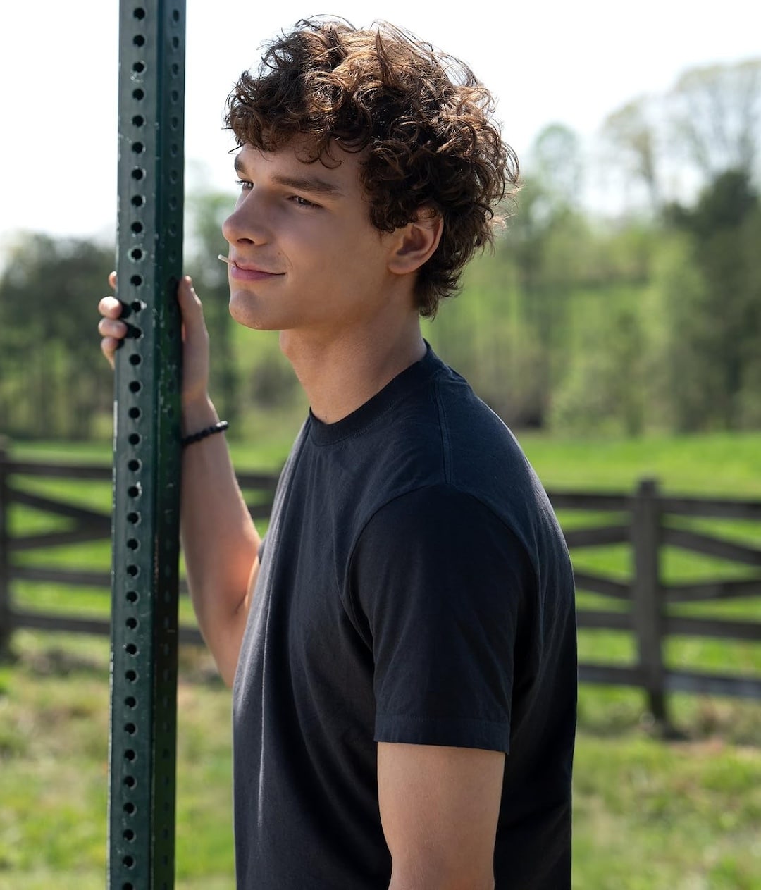 A young man with curly brown hair, wearing a black t-shirt, stands outdoors by a metal post, with a wooden fence and green trees in the background. Reminiscent of Conrad Fisher, he is looking slightly to the side and smiling gently.