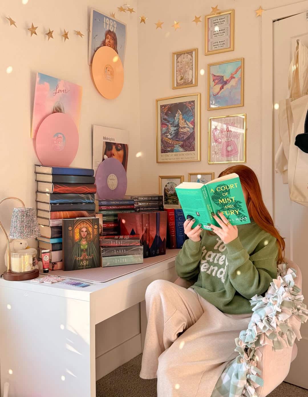 A person with long hair sits at a desk in a cozy room, reading A Court of Mist and Fury. The desk holds books, icons, and decorations; the wall behind features vinyl records, framed art, and string lights.