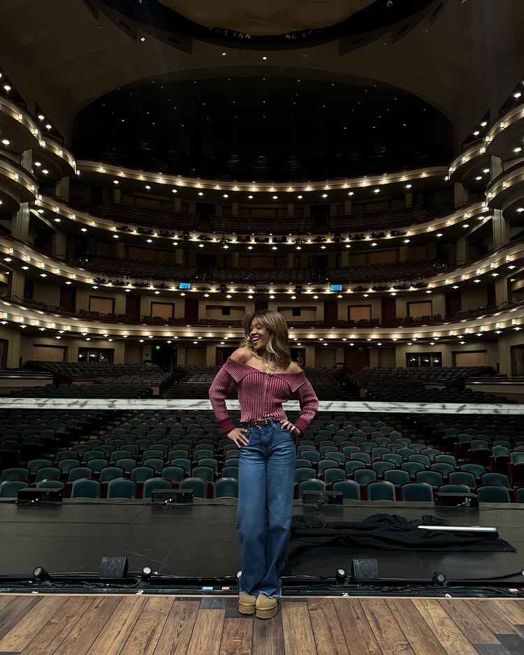 A woman in a maroon off-shoulder sweater and blue jeans stands confidently on a theater stage, smiling, ready to ace auditions, with empty seats and ornate balconies in the background.