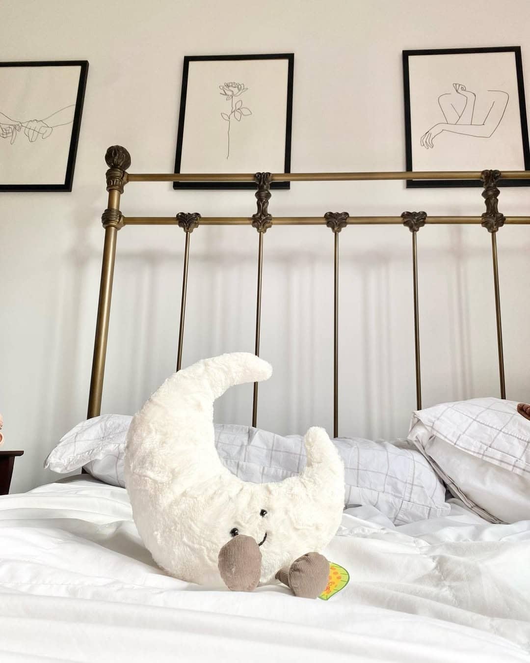 A plush crescent moon toy sits on a neatly made white bed with pillows. Behind the bed, a brass headboard and three minimalist line art drawings in black frames create a calming space perfect for reflecting on your moon sign or favorite astrology insights.