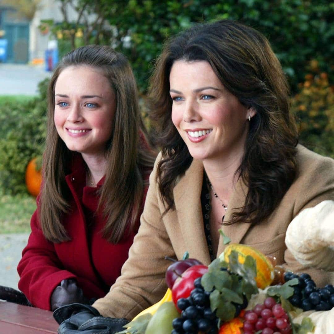 Two women with long brown hair sit side by side at a table outdoors, smiling. Reminiscent of fictional mother-daughter duos, one wears a red coat and gloves, the other a tan coat. A colorful arrangement of fruit and vegetables sits before them.