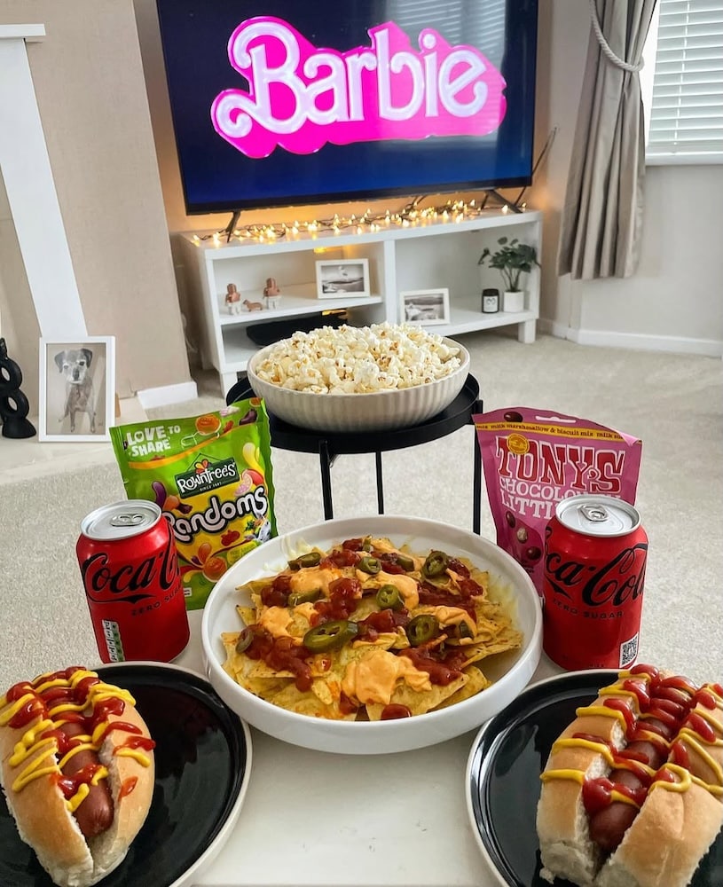 A cozy living room setup perfect for winter activities, with a TV showing the Barbie logo. On the table are two hot dogs, nachos with toppings, popcorn, Coca-Cola cans, candy, and chocolate—ideal for a friend hangout and movie night when it’s cold out.