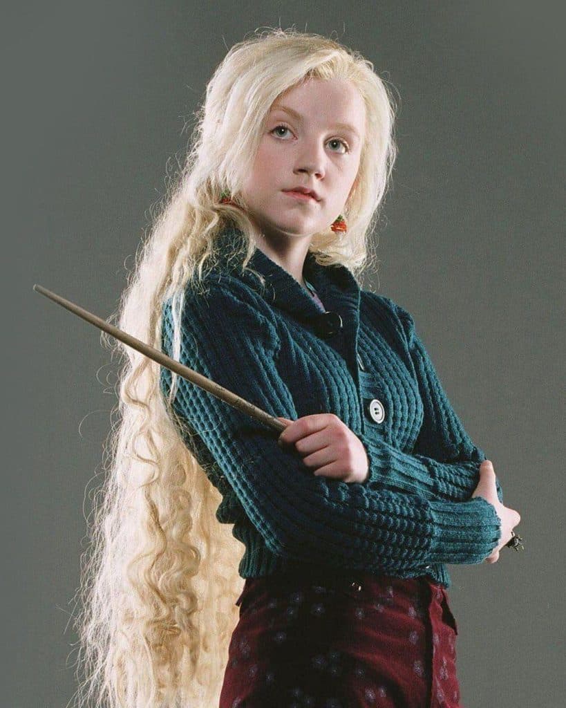 A young person with very long, wavy blonde hair holds a wand and looks confidently at the camera. They wear a dark teal sweater and maroon skirt, standing against a plain gray background.