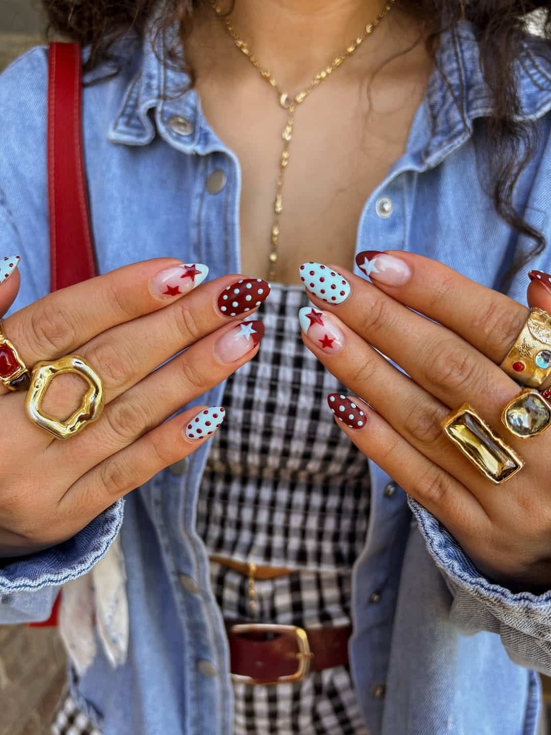 Close-up of hands with long, almond-shaped nails featuring red and blue polka dots and star designs—a nod to 2026 nail trends. Chunky gold rings, a denim jacket, and a black-and-white checkered dress complete the look.