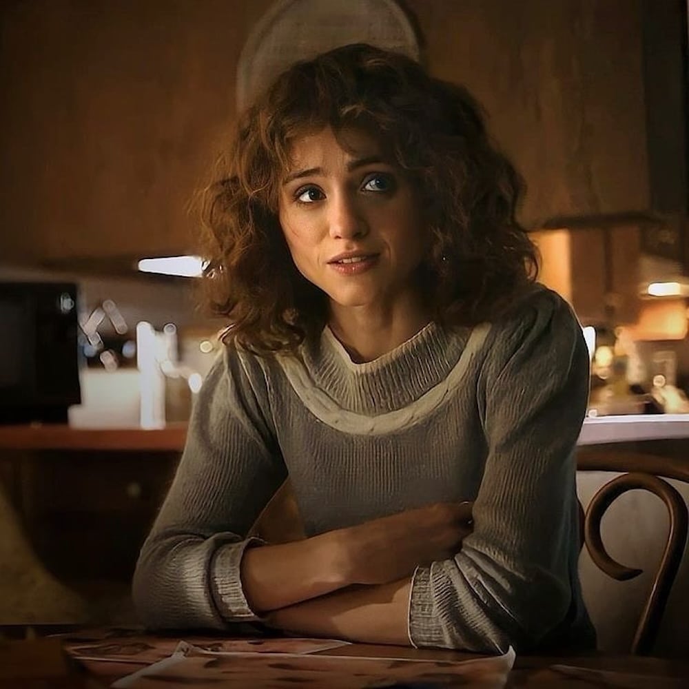A woman with curly brown hair and a gray sweater sits at a kitchen table, looking slightly concerned. Warm lighting creates a cozy atmosphere, as if she could be a character from Stranger Things, with kitchen items visible in the background.