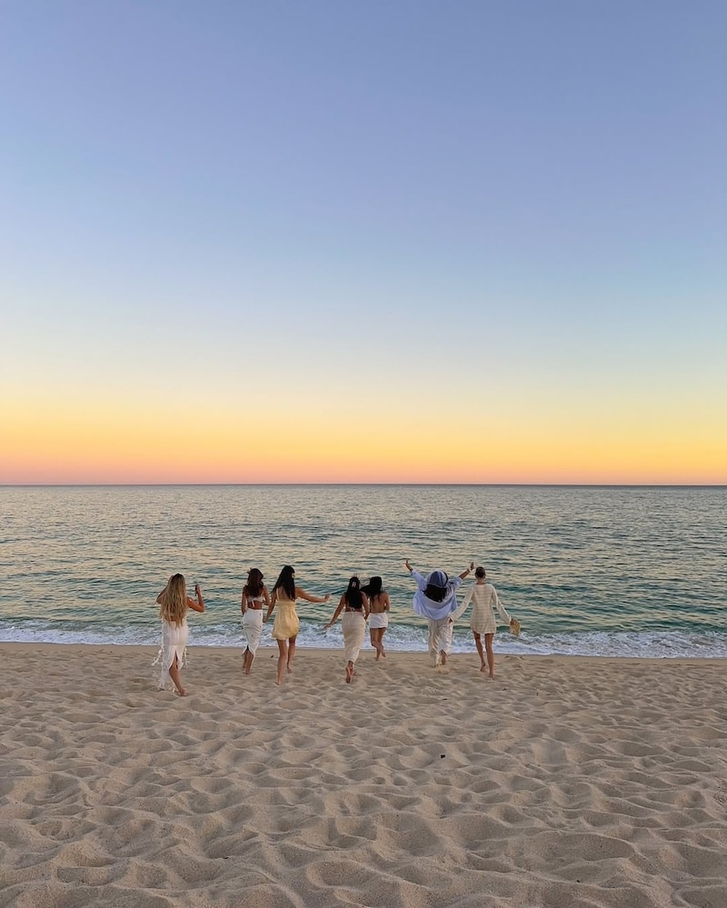 Six people in light clothing walk toward the ocean on a sandy beach at sunset, with a colorful sky fading from yellow to blue above calm waves.