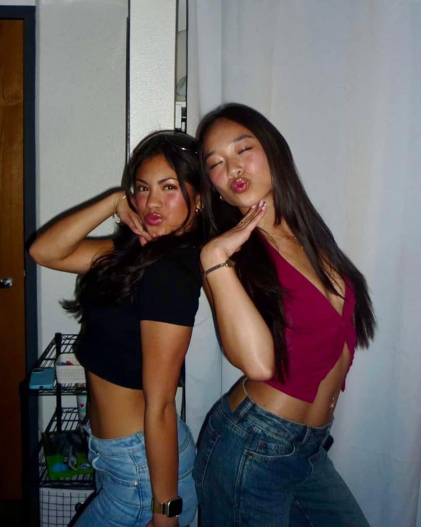 Two young women pose playfully, standing side by side with one hand under their chins and lips puckered. Sporting casual crop tops and jeans, they radiate Galentine’s Day hype in a brightly lit indoor setting.