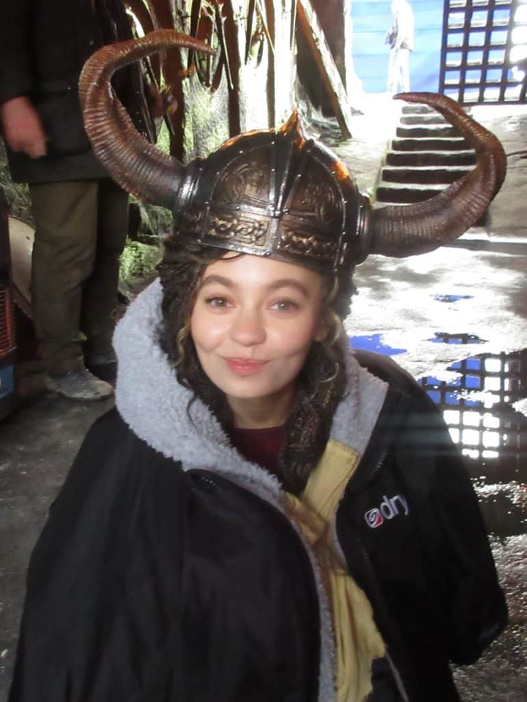 A young woman smiles at the camera while wearing a Viking-style horned helmet and a large black jacket, standing in a dimly lit area with wet ground and metal bars in the background.
