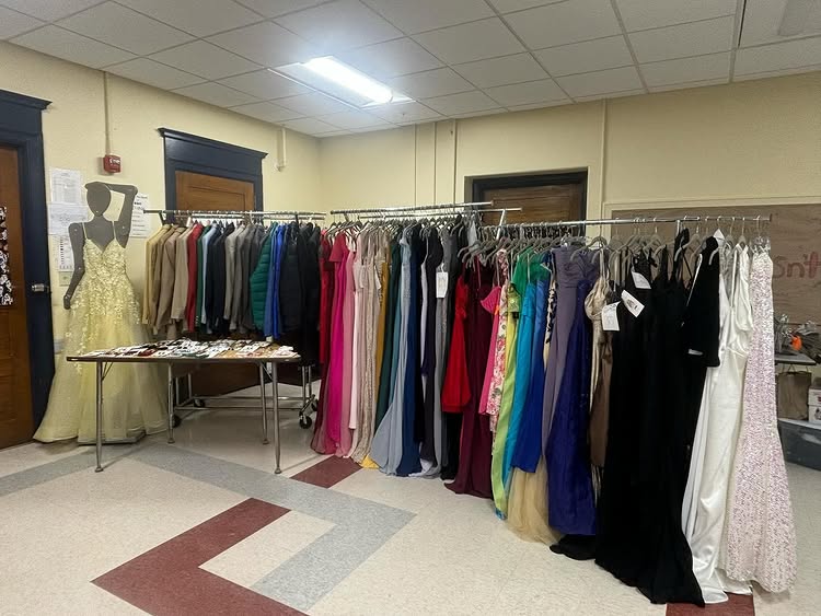 A room with racks of colorful formal dresses and suits, a table displaying jewelry, and a mannequin in a yellow gown highlights Ocean Lombard’s commitment to sustainable fashion, ideal for Miss Rhode Island's Teen or prom events.
