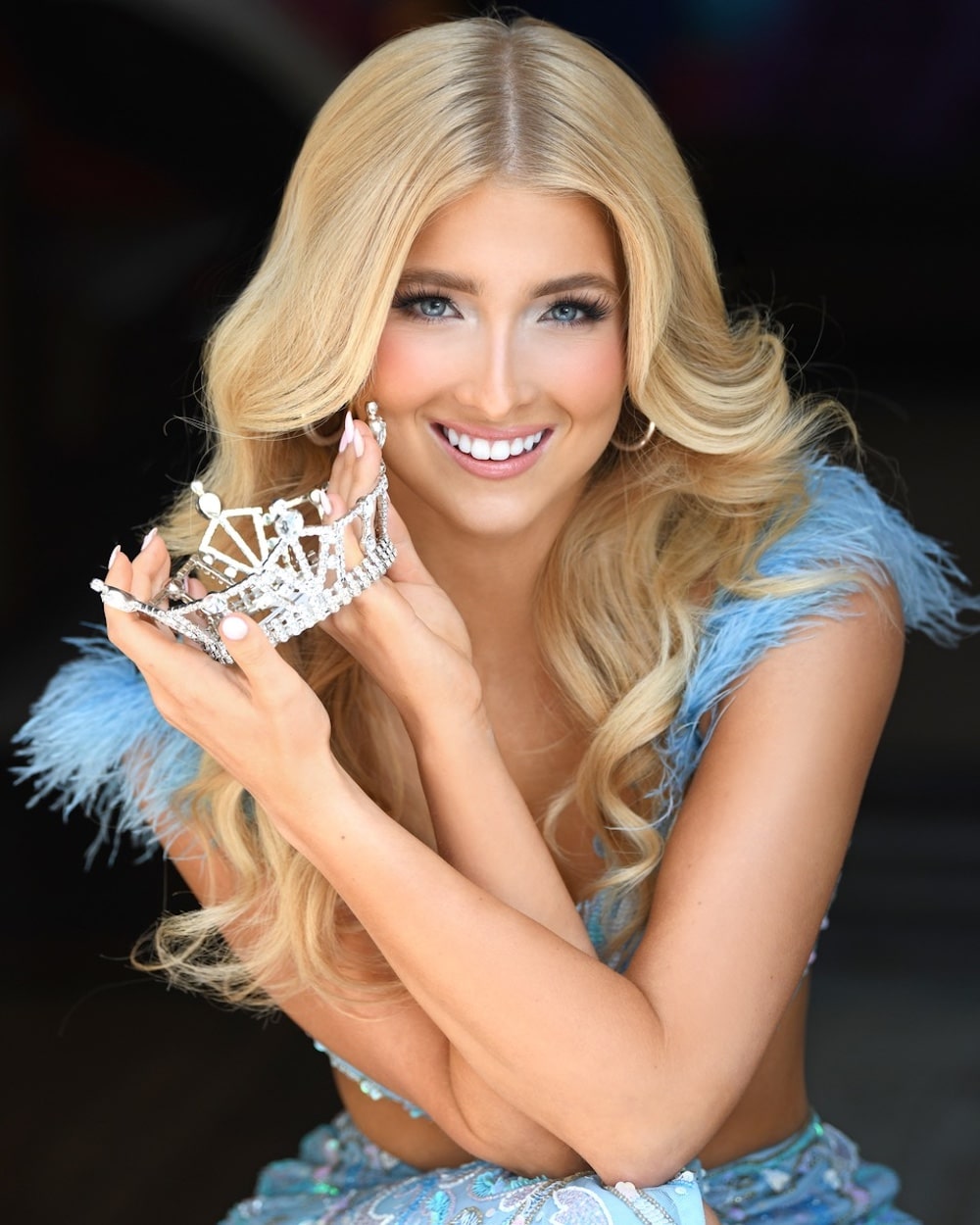 A smiling woman with long blonde hair holds a sparkling crown near her face. She wears a light blue, feather-trimmed dress and has manicured nails, posing against a dark background.