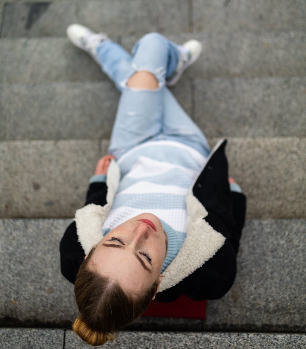 A person with light brown hair in a bun, wearing a light blue sweater, jeans, and a black jacket with a shearling collar, sits on stone steps with eyes closed—perhaps recalling awkward moments or an embarrassing situation with a crush.