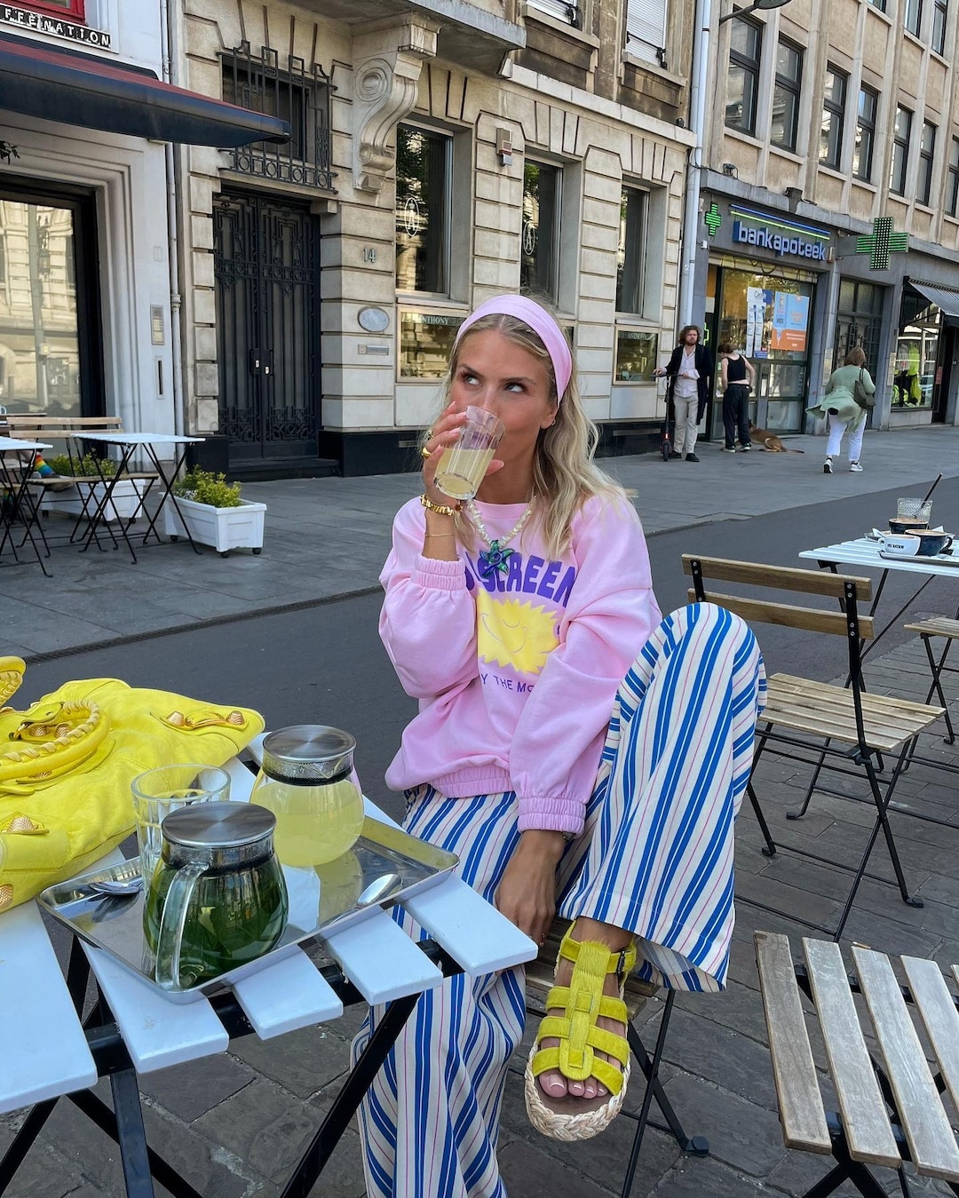 A woman enjoying a spring refresh sits at an outdoor café table in a pink sweatshirt, striped pants, and yellow sandals—an inspiring closet update with city streets and buildings visible in the background.