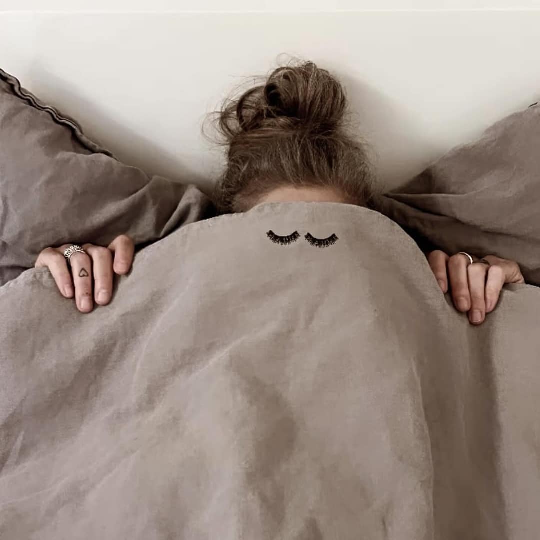 A person lies in bed mostly covered by a gray blanket, with only their hands and messy hair visible. The blanket, perfect for period pain relief, has small, closed-eye illustrations near the top.