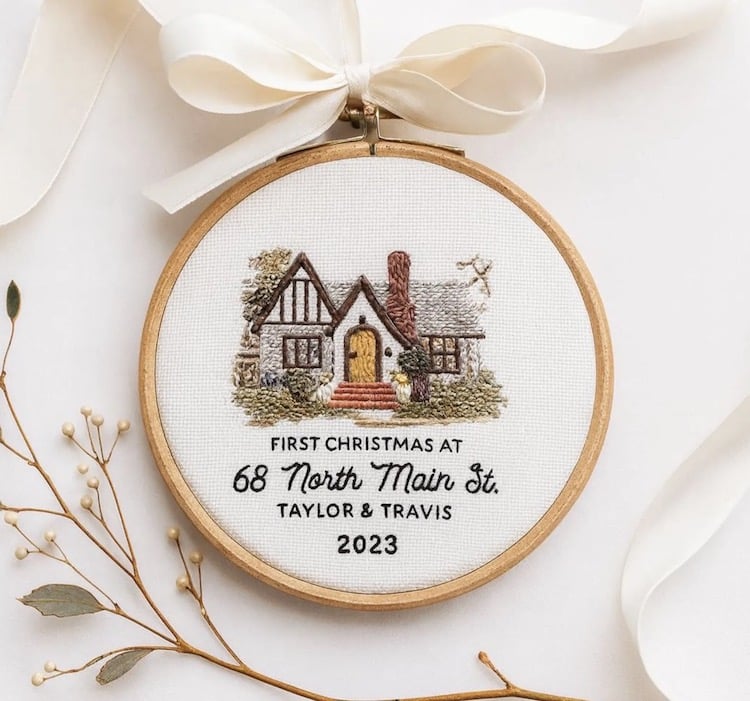 An embroidery hoop displays a cozy house with trees and a path, with text below that reads: “First Christmas at 68 North Main St. Taylor & Travis 2023.” A white ribbon and small dried flowers decorate the scene.