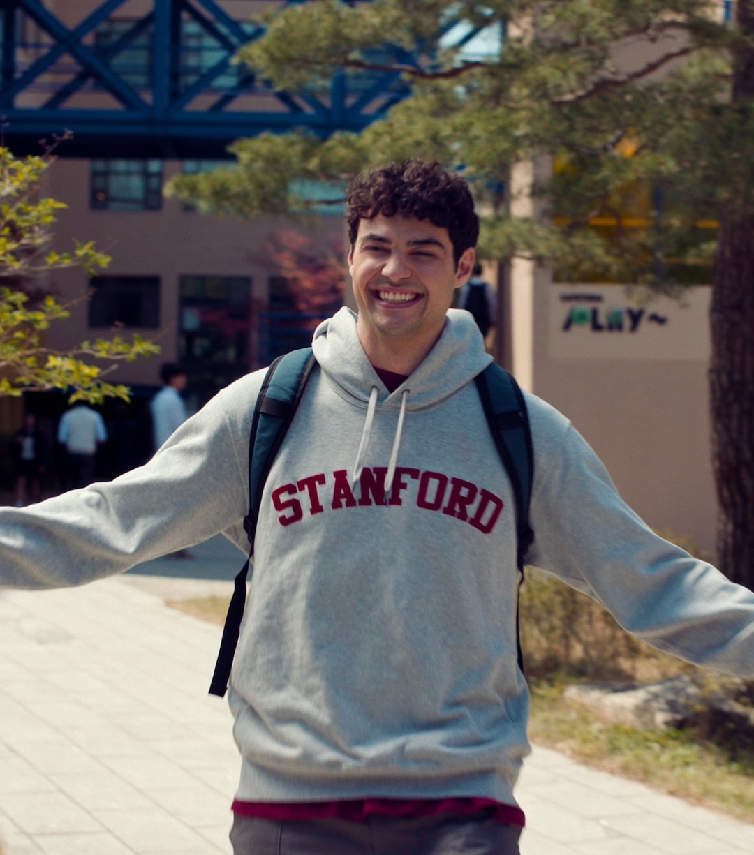 A smiling young man, Conrad Fisher, wearing a gray Stanford hoodie and backpack stands outdoors with arms open, in front of a building and trees on a sunny Valentine’s Day.