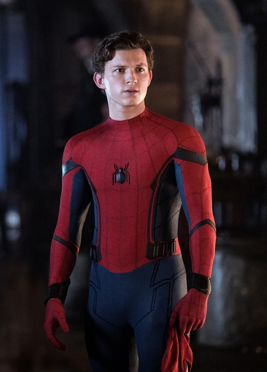 A young man in a red and blue Spider-Man suit stands indoors, holding part of his mask in one hand with a serious expression—reminiscent of Conrad Fisher on Valentine's Day—with a blurred background behind him.