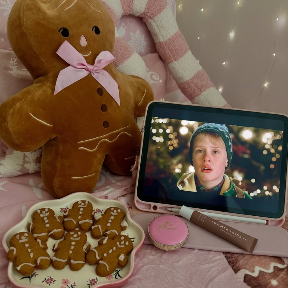 A cozy Pink Peppermint Christmas scene with a gingerbread man pillow, a plate of gingerbread cookies, lip gloss, a pink container, and an iPad showing the young boy from Home Alone—all on a pink bedspread.