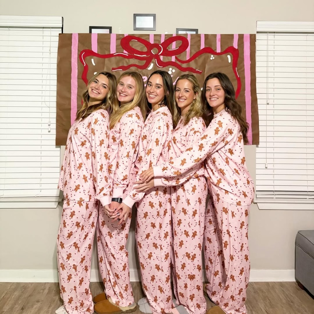Five women, all BFFs, stand side by side in matching pink gingerbread pajamas, smiling in front of a festive gingerbread house. The cozy room with white blinds sets the perfect scene for an aesthetic Christmas sleepover.