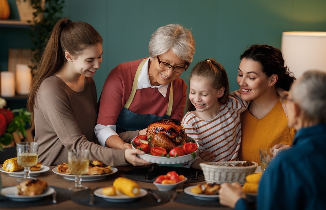 A smiling older woman serves a roasted turkey to three happy family members at a dinner table, where laughter and light conversation—perhaps even talk politics—fill the warmly lit, cozy dining room.