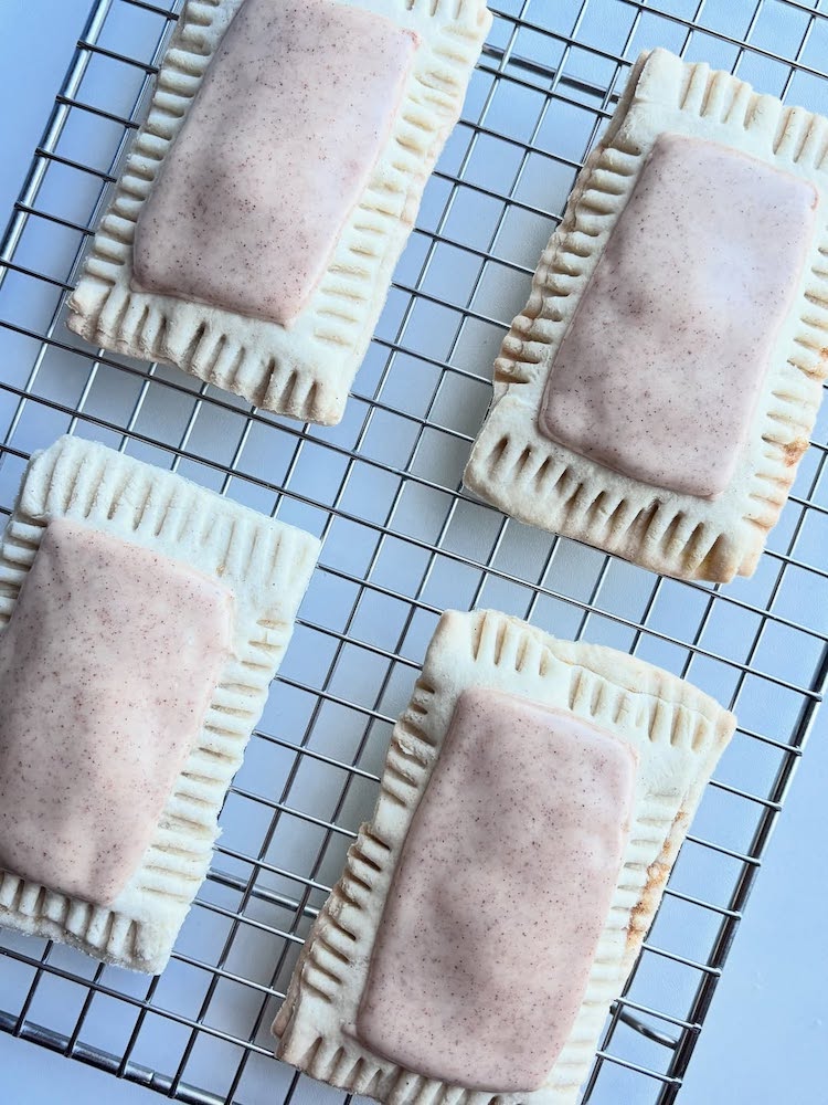 Four rectangular pastries with light brown frosted tops and crimped edges are cooling on a wire rack against a white background.