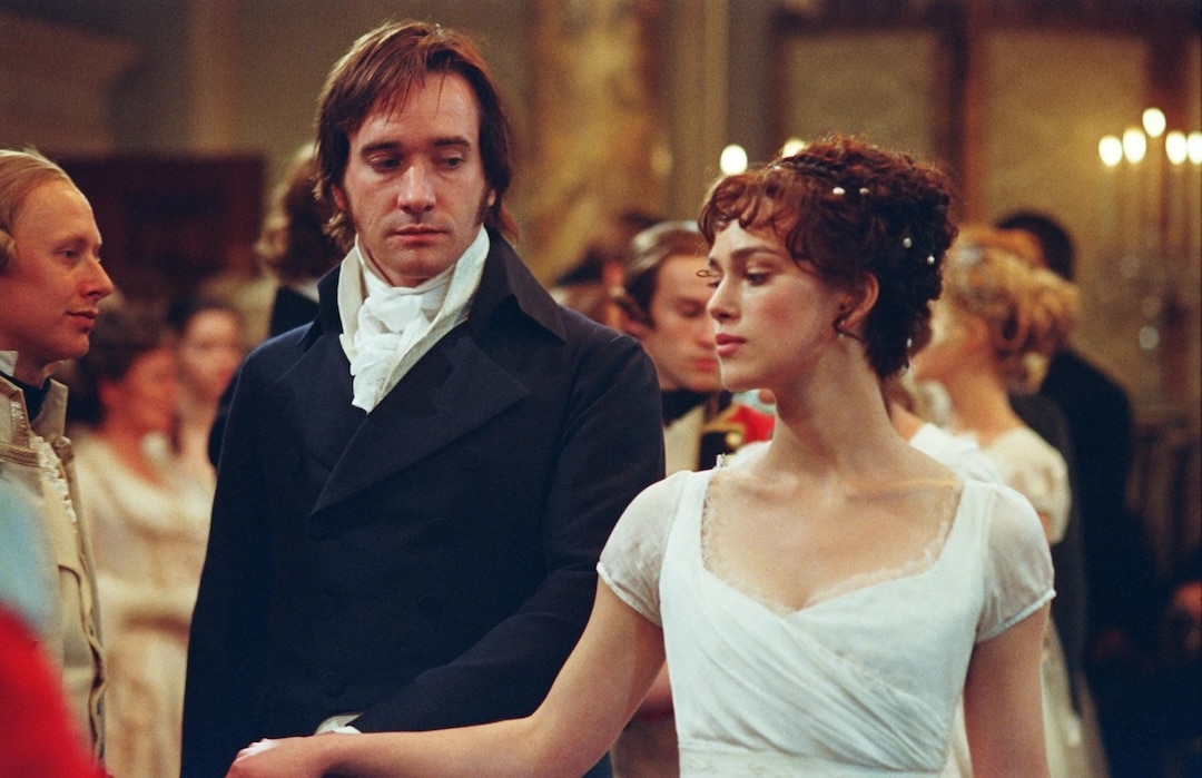 A man in a dark suit and a woman in a white dress stand close together at a formal ball, as if stepping out of one of the most perfect book-to-movie adaptations, surrounded by elegantly dressed guests in a grand room with soft lighting.