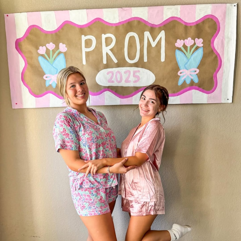 Two young women in pajamas smile and pose holding hands in front of a handmade PROM 2025 sign with pink tulips. Against a neutral wall, they look happy and excited—perfect inspiration for your own BFF brunch or prom brunch ideas!.