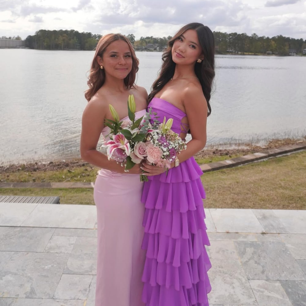 Two women stand side by side by a lake. One wears a light pink dress and holds a bouquet of flowers; the other wears a strapless, ruffled purple dress. Both are smiling, and the background shows trees and water under a cloudy sky.