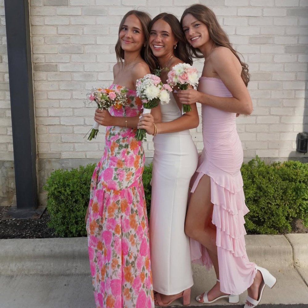 Three young women in colorful formal dresses hold small bouquets of flowers and pose together, smiling, in front of a light brick wall and green shrubs.