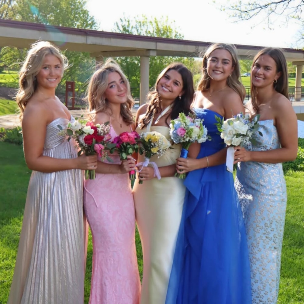 Five young women in colorful formal dresses stand outside on grass, holding bouquets and smiling at the camera. The background features trees and a building on a sunny day.