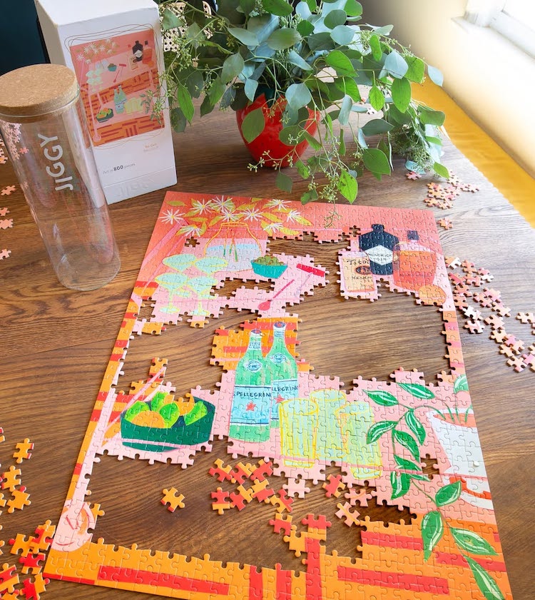 A colorful jigsaw puzzle depicting drinks, fruit, and plants is partially completed on a wooden table. Puzzle pieces, a glass water bottle labeled Jiggy, and a leafy potted plant are nearby in bright natural light.