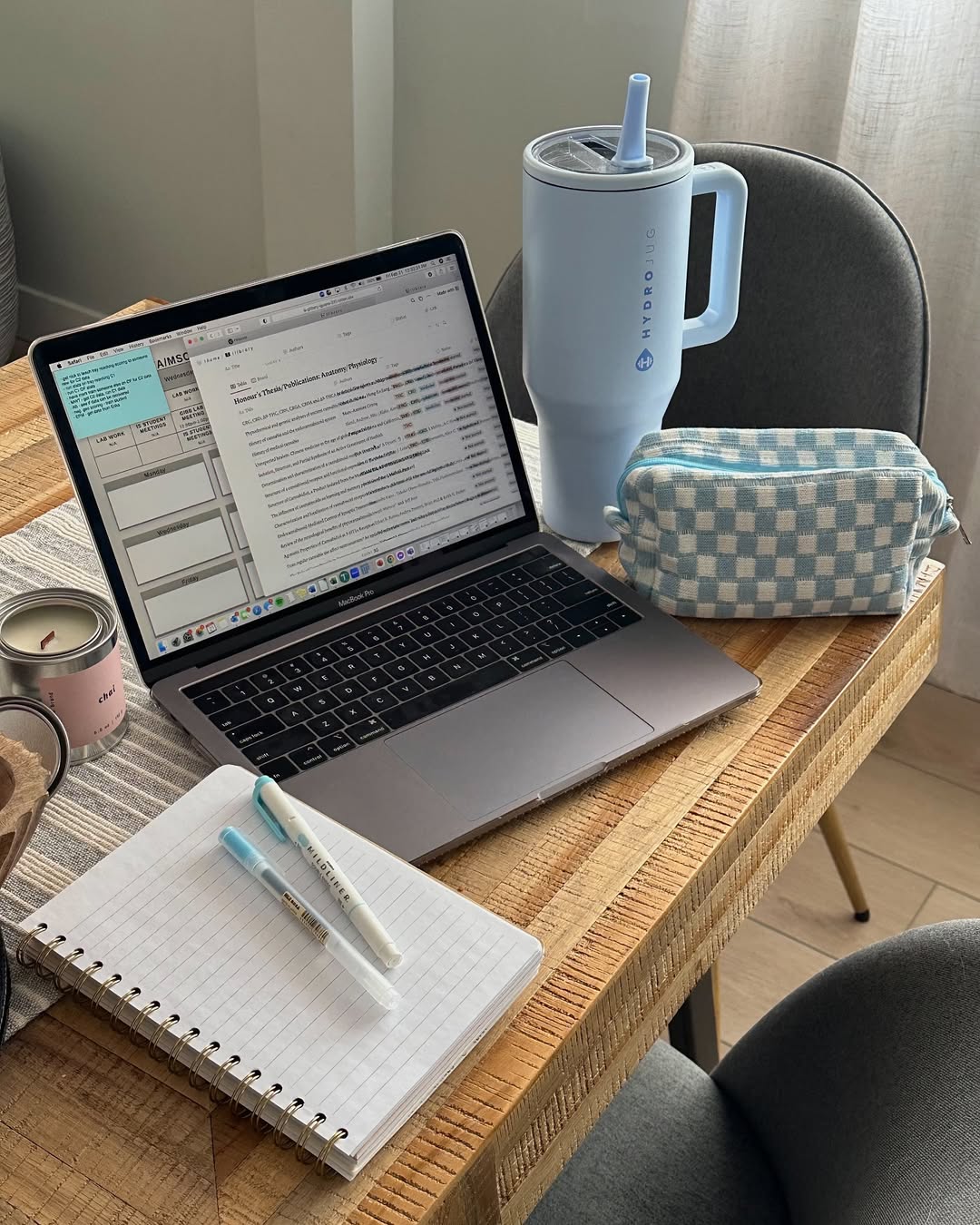 A laptop on a wooden table with an open planner, two pens, a notepad, a blue checkered pouch, a large light blue tumbler, and a lit candle creates a cozy workspace by a window—perfect for jotting down your favorite ChatGPT tips.