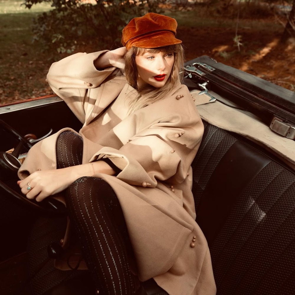 A woman wearing a tan coat, black patterned tights, and a rust-colored cap sits in the driver’s seat of a vintage convertible car, looking to the side with red lipstick and autumnal surroundings in the background.