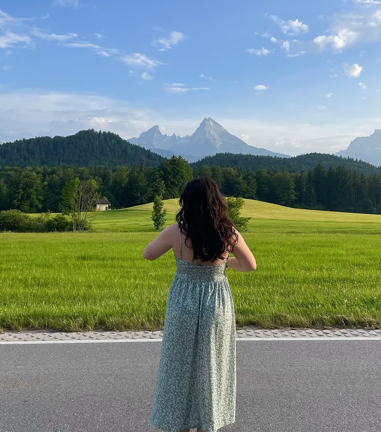 A woman in a light green dress stands on the roadside facing green fields and mountains under a blue sky. With her back to the camera, she enjoys nature—a reminder to reduce screen time and embrace the beauty around us.