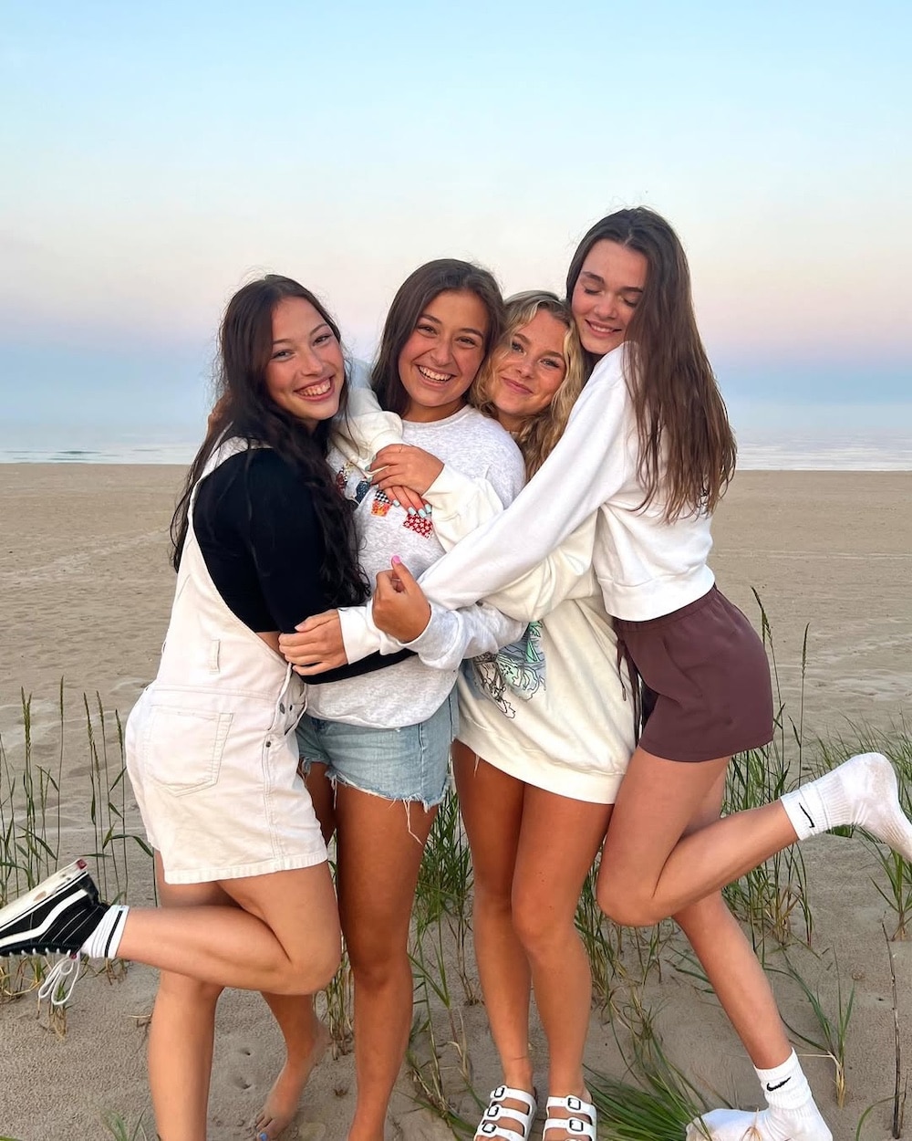 Four young women stand together on a sandy beach at sunset, smiling and hugging each other. Dressed in casual summer clothes, they radiate joy, reminding us to stop comparing and simply love life with close relationships.