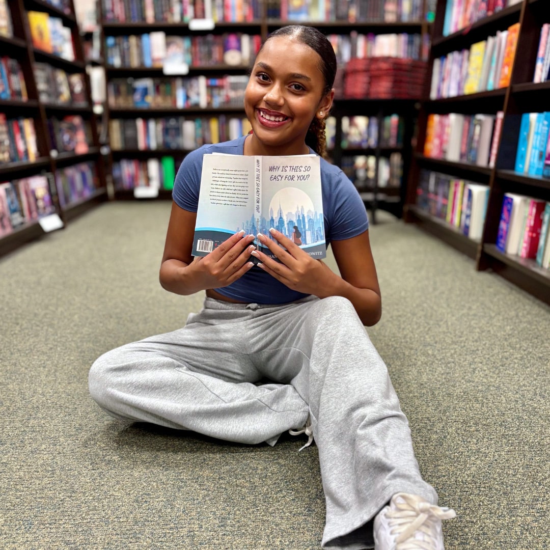 A young woman sits cross-legged on the floor of a bookstore, smiling and holding an open book, surrounded by colorful shelves—finding balance between juggling school and her social life.
