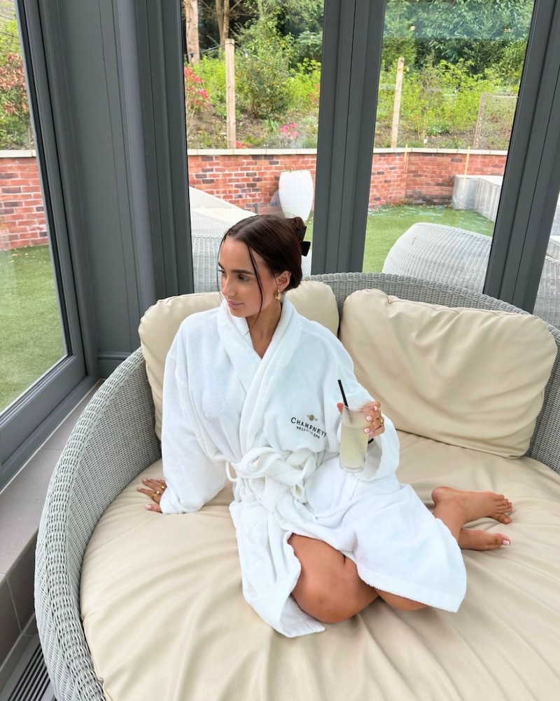 A woman in a white robe sits on a round cushioned sofa in a bright sunroom, enjoying an at-home spa day with a drink as she looks out the window at the lush green lawn and trees.
