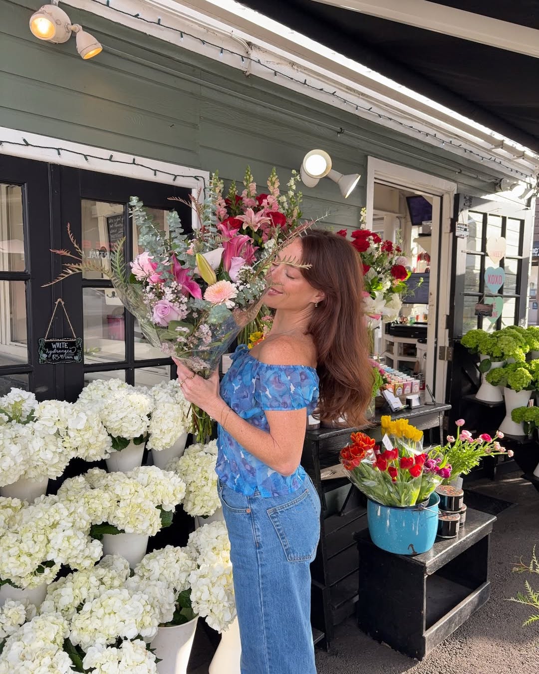 A woman in a blue off-shoulder top and jeans holds a large bouquet of flowers, smiling with her eyes closed, inviting you to romanticize your spring as she stands before a flower shop filled with vibrant blooms.