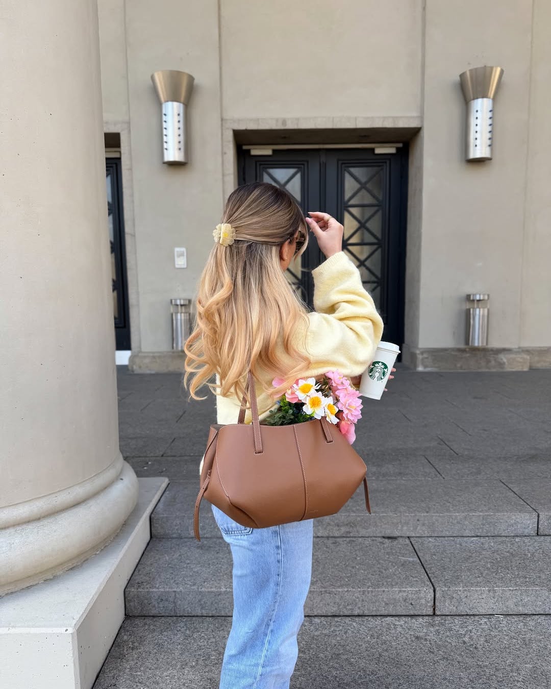 A woman with long, wavy, blonde hair stands outside a building holding a Starbucks cup. Dressed in spring-inspired colors, she carries a brown handbag with colorful flowers peeking out—perfect to romanticize spring and make life less boring.