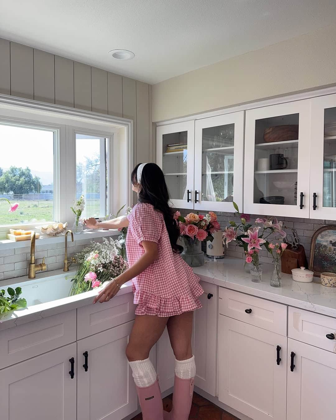 A woman in a pink dress and boots arranges flowers by a kitchen sink, with sunlight streaming through the window—a perfect way to romanticize your spring, surrounded by vases of fresh flowers on the white countertop.