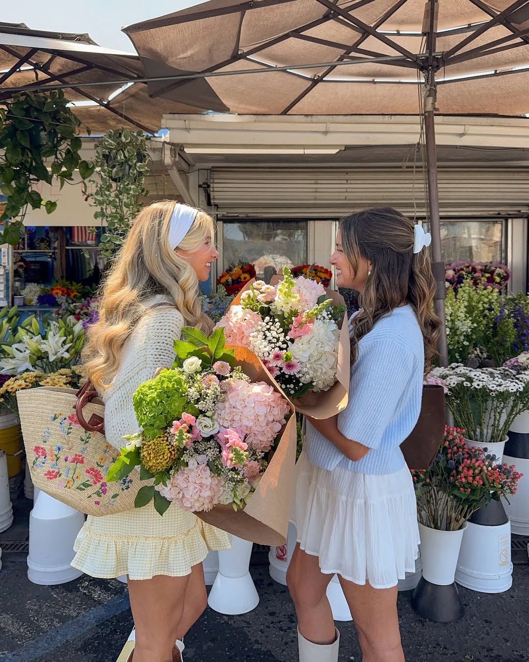Two women stand outdoors at a flower market, smiling at each other while holding large bouquets of colorful flowers. Dressed in light spring outfits, they embody spring inspiration as they romanticize life among vibrant arrangements in buckets behind them.