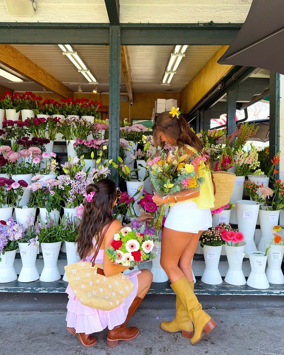 Two women in colorful outfits, surrounded by vibrant blooms and white vases, pick flowers at an outdoor market—capturing the essence of spring romance and inspiring all to romanticize your spring with every bouquet.