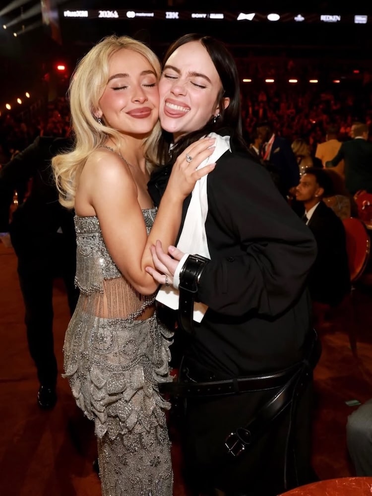 Two young women smile and hug each other at an indoor event, capturing the joy of unforgettable Grammy moments. One wears a sparkly silver outfit, the other wears black, with seated guests and bright stage lights in the background.