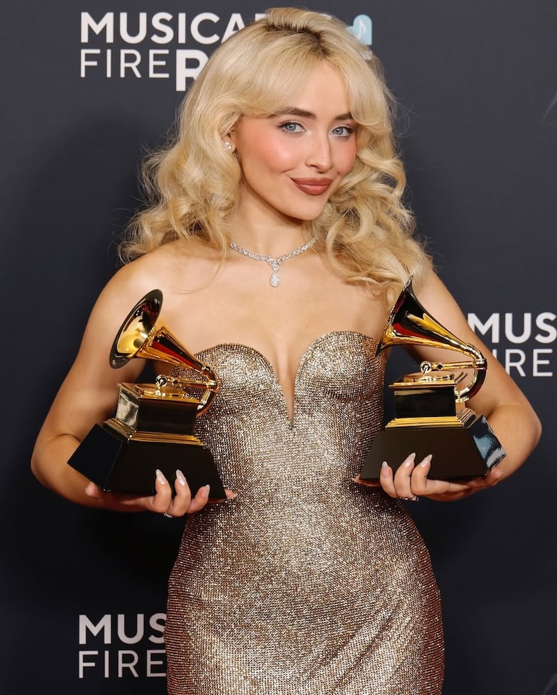 A woman with long blonde hair wearing a sparkling strapless gown holds two Grammy awards, standing in front of a black backdrop with MUSICAL FIRE written on it. She is smiling slightly, showing that sometimes Dreams Come True for Disney Stars.