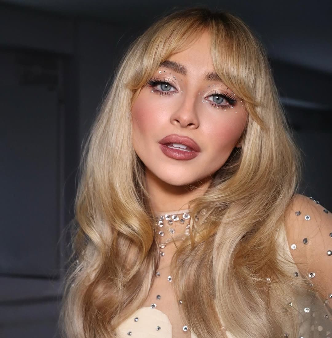 A woman with long, wavy blonde hair and bangs poses indoors, decked out in sparkly makeup with rhinestones by her eyes and a sheer, pearl-embellished top. The softly blurred background hints at festive Christmas decor.