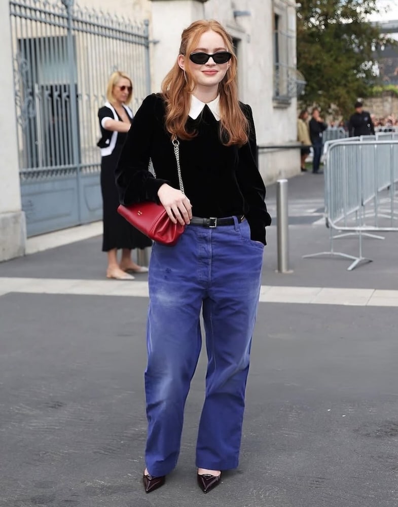 A woman with long red hair, wearing black sunglasses, a black sweater with a white collar, loose blue pants, maroon heels, and carrying a red quilted purse, stands outdoors near metal railings and a gate.