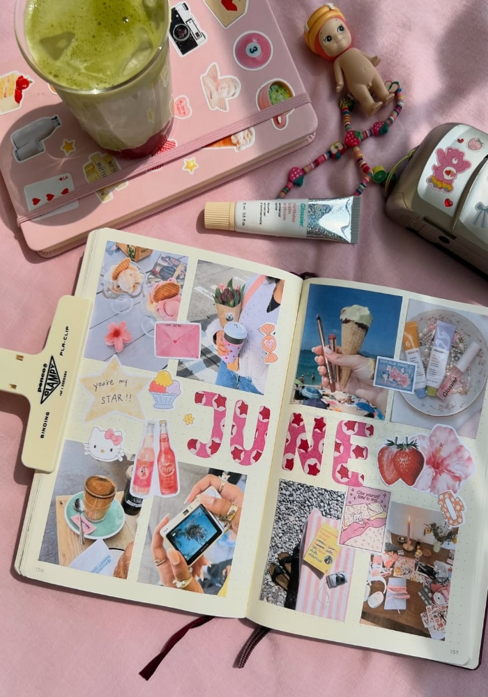 A scrapbook journal open to a June-themed collage page with photos, stickers, and colorful decorations—perfect for BTS scrapbook inspiration—surrounded by a matcha drink, toy doll, decorated notebook, camera, and lip balm on a pink cloth.