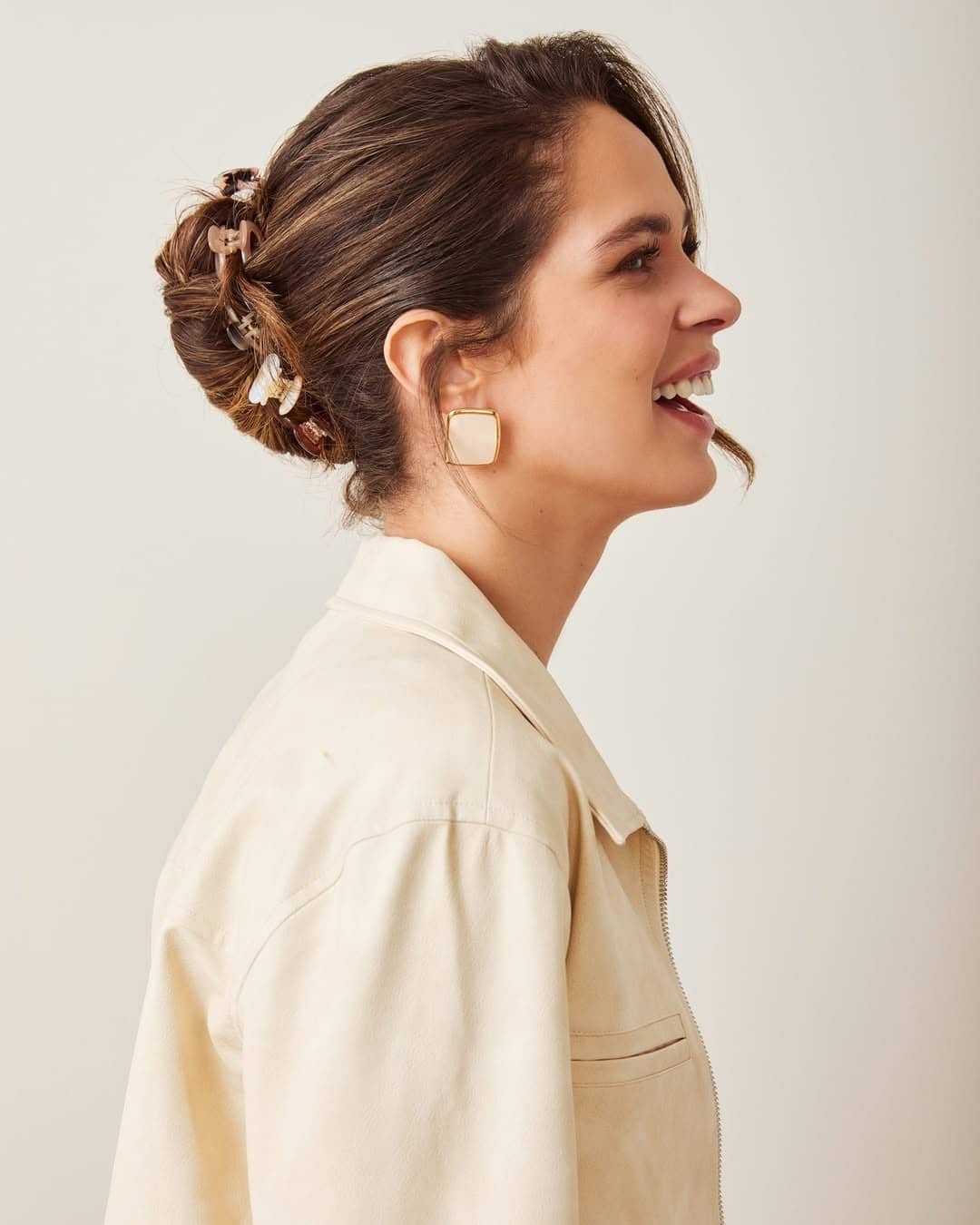 A woman with brown hair styled in a bun, secured with a chic claw clip and other hair accessories, wears a light beige jacket and large square earrings, smiling while looking to the side against a plain background.