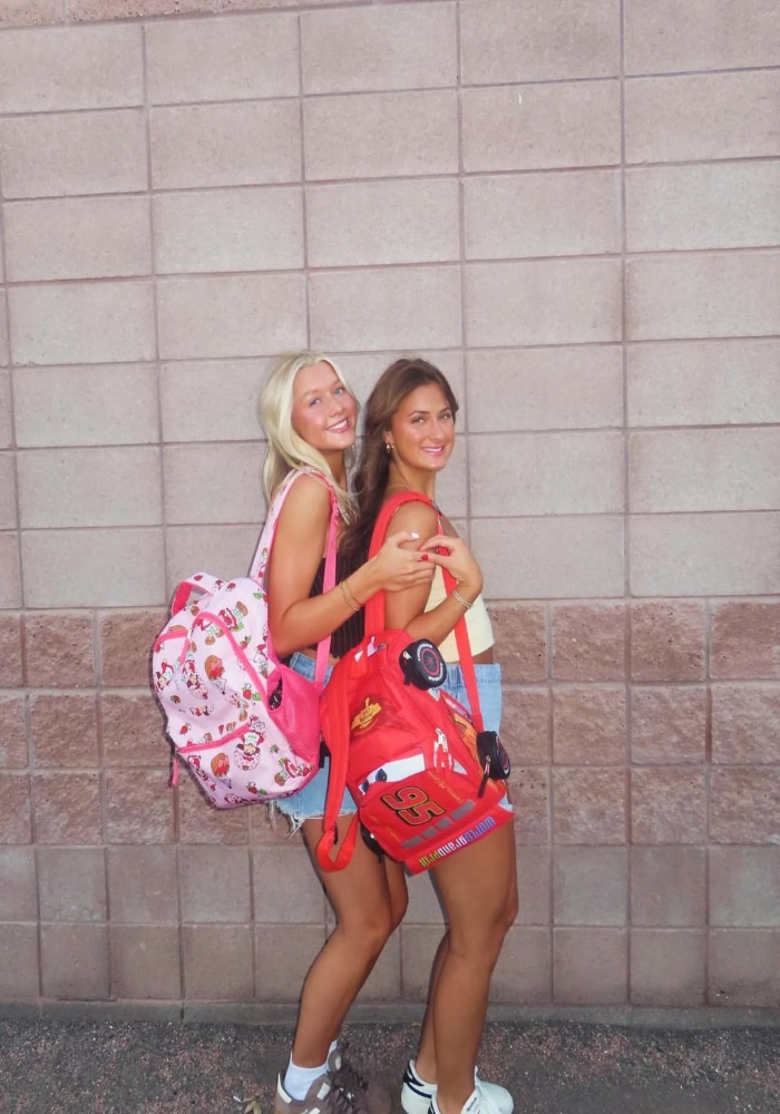 Two young women stand smiling in front of a brick wall, each carrying colorful childrens backpacks—one pink with hearts and the other red with a racecar design. They are dressed casually and appear cheerful.