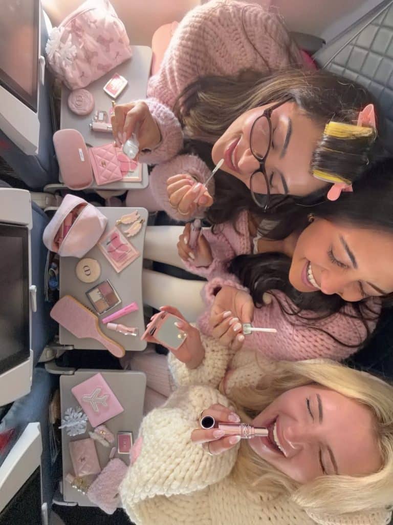 Three women sitting on an airplane, smiling and applying makeup with various pink cosmetic products spread out on their tray tables, enjoy a cozy Galentine’s Day together in their sweaters.